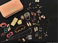 Photo displaying pink velvet jewelry case and assorted costume jewelry including earrings, necklaces, brooches, and bracelets on a black surface.