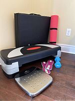 Overview photo showing the aerobic step platform, folded black exercise mat, pink yoga mat, blue dumbbell, wrist weights and digital scale all arranged on floor.