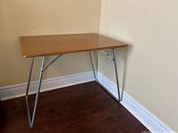 Retro folding table with a rectangular wood veneer top and tubular metal legs, positioned in corner against walls on a dark wood floor.
