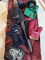 Photo shows a burgundy travel golf bag, black wrapped golf umbrella, Ping black bucket hat, red Ping mitt, navy Sleepy Hollow mitt, and a green plastic putting cup along with a wooden driver headcover.