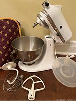 White KitchenAid mixer with stainless bowl, three attachments, plastic lid, and dust cover.