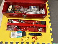 Photo showing the contents of the open red metal tool box on a yellow foam mat. Items include electrical outlets, screws, wires, adapter plugs, and testers.