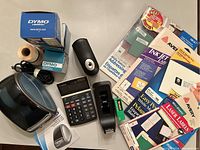 Full view of lot items on tabletop showing Dymo LabelWriter in box, calculator, pencil sharpener, tape dispenser, and label packages.