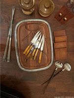 Wooden steak board with six steak knives, four coasters, four skewers, salt and pepper mill, vintage salad servers, and two crocks arranged on a wooden surface.