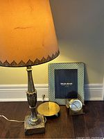 Photo showing vintage brass lamp with off-white worn shade, solid brass picture frame with striped border, brass pillar candle holder, and Tiffany brass round clock with square base.