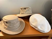 Set of three hats on wooden surface: two beige Tilley hats and one white Kangol golf cap.