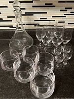 Photo of a crystal glass decanter with stopper, six cocktail glasses, seven sherry glasses, and four aperitif glasses arranged on a countertop with a tile backsplash.