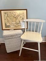 Photo showing the white wooden chair, wicker hamper with lid, and vintage framed map together against a pale blue wall.
