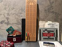 Photo of wooden cribbage board standing upright at center with pegs, Auto Bridge game tray with dice and instruction papers, two deck card boxes with decorative card backs and suits displayed.