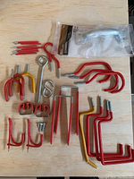 Assorted red and yellow painted metal hooks, various sizes and shapes with screw ends, metal eye hooks, S-shaped hook, and a packaged silver door pull handle on wood surface.