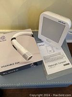 Photo showing the Verilux HappyLight Compact UV therapy light alongside its manual and the Blaux Wearable Fan box on a blue surface.