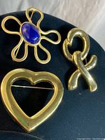 Three gold tone brooches on black display; flower with blue stone, XO symbol, hollow heart shape.