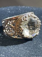 Close-up photo of ornate sterling silver ring with clear faceted stone and detailed filigree band, showing front and side profile.