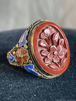Close-up image showing the red cinnabar wood oval top of the ring with floral carving and metal detailing on the band with blue and orange stones.