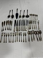 Overview of the flatware set laid out, showing spoons, forks, knives, and serving utensils arranged on a white cloth.