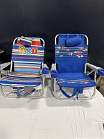 Front view of two beach chairs side by side on white surface against black background, showing fabric patterns and cooler pouches