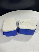 Two blue plastic coolers with white lids closed, tops dirty and marked, placed on white fabric surface.