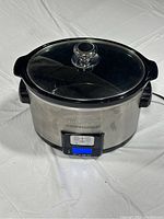 Full view of stainless steel programmable slow cooker with glass lid on a white surface.