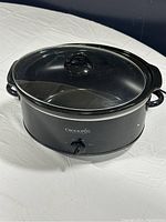 Black oval manual slow cooker with glass lid on white surface, shows full device from top angle