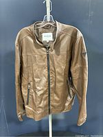 Front view of Guess Los Angeles brown faux leather jacket hung on a hanger against a dark background showing the full front and zip details.