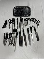 Full view of all 25 kitchen cutting utensils including knives, scissors, scoops, whisk, ladle, spatula, strainer, and black serving tray.