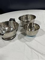 Three stainless steel bowls on white cloth: one colander with handles and two mixing bowls.