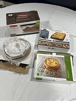 Four kitchen bakeware items displayed on white surface. One white round baking pan in packaging, one baking tray box with fries on cover, and two boxed metal cooling racks.