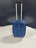 Full frontal image of blue luggage suitcase with extended handle and wheels visible, background grayish, on a white cloth surface