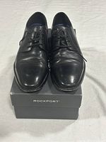 Front view of black leather men's dress shoes on Rockport box showing laces, toe shape, and condition.