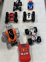 Full lot photo of six toy cars and vehicles arranged on white cloth background, showing large wheels and various colors.