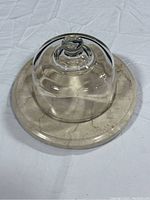 Clear glass dome with knob handle placed on beige round marble base showing natural veining, photographed from top view.