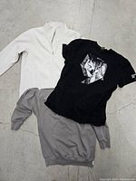 Three men's tops including white zip-up jacket, black graphic tee, and gray long sleeve shirt laid flat on concrete floor