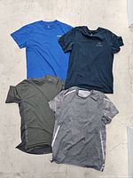 Four men's medium shirts laid flat on concrete floor, showing front side and logos in color black, blue, olive green, and gray.