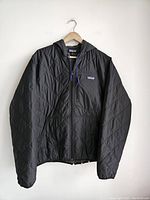 Front view of black quilted jacket with hood, hanging on wooden hanger against white wall