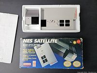 Remote control module in white with black buttons inside original styrofoam packaging. Box front with NES Satellite branding and turbo control features visible
