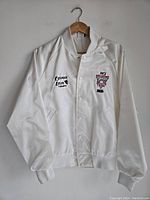 Full view of white satin sports jacket showing snap button front, ribbed cuffs, and collar with embroidered logos on chest.