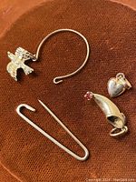 All five gold pieces arranged on brown background: hoop earring with bird, shoe and heart charms, bar pin and stick pin