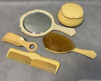 Vanity set items arranged on a surface: hand mirror, large powder box with lid, hairbrush, and two combs.