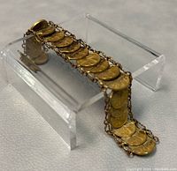 Side angle of bracelet resting on a clear plastic stand showing linked Mexican coins with chain links and clasp closure.