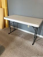 White rectangular folding table with gray metal legs positioned against a wall, showing the tabletop and legs.