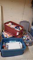 Four Rubbermaid totes filled with assorted acrylic paints, brushes, canvases, and art supplies viewed together.