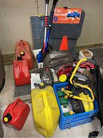 Photo showing car floor mats, multiple gas cans, booster cables, trouble light, snow brushes, and safety cone grouped on floor.