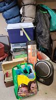 Full view of various camping and outdoor items stacked, including coolers, sleeping bags, wooden box, and a Coleman heater.