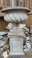 Full side view of concrete planter showing twisted design and pedestal base, surrounded by stones and wood pieces.