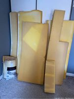 Multiple yellow foam pieces of various sizes and thickness approximately 4 inches, standing upright against a wall beside a packaged mattress pad.
