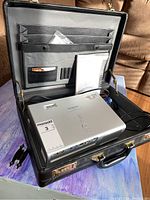 Hitachi projector inside an open black briefcase showing cables and user manual.