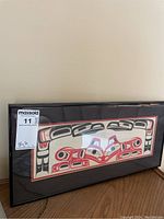 Single black framed Inuit art print with red and black design, lying on a wooden surface, showing the full piece horizontally.