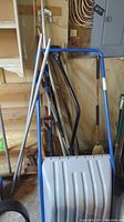 Tools and snow shovels leaning against garage wall including plastic snow shovel, metal shovels, blue snow rake, broom, rake, and brush