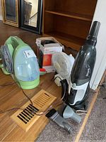 Lot items on floor near bookshelf: green Bissell Little Green shampooer, white oscillating ceramic heater box behind small white table fan, and black Shark hand vacuum with attachments in front.
