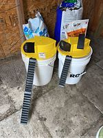 Two white plastic buckets with large yellow mouse trap lids and black ramps attached, placed on a concrete floor in a wooden shed environment, with fertilizer and soil bags in background.